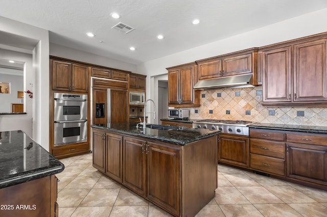 kitchen with visible vents, an island with sink, a sink, under cabinet range hood, and built in appliances