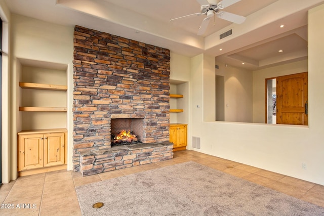 unfurnished living room featuring built in features, ceiling fan, light tile patterned flooring, a stone fireplace, and a raised ceiling