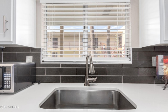 kitchen view of a sink, stainless steel microwave, white cabinetry, and backsplash