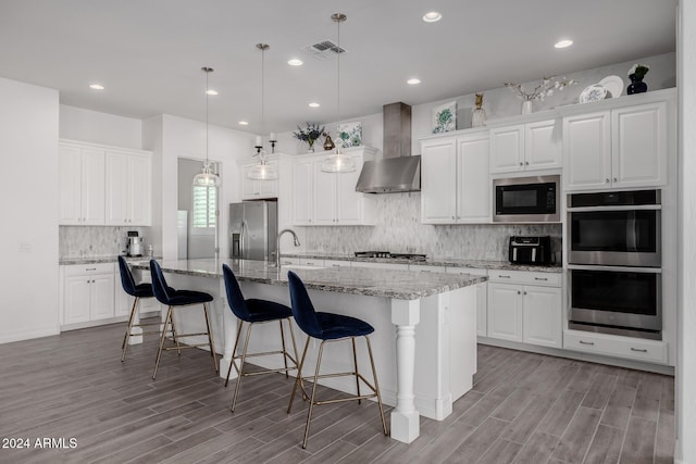 kitchen with stainless steel appliances, white cabinetry, wall chimney exhaust hood, and an island with sink