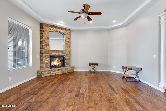 unfurnished living room with a stone fireplace, hardwood / wood-style floors, decorative columns, a tray ceiling, and ceiling fan