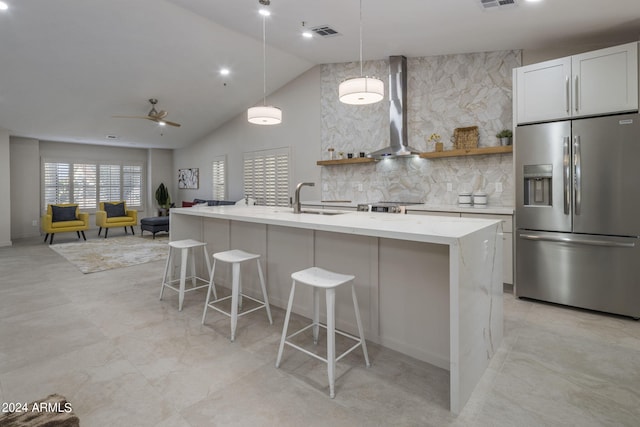 kitchen with an island with sink, hanging light fixtures, stainless steel appliances, and wall chimney range hood