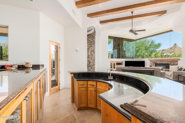 kitchen with dark stone counters, ceiling fan, sink, beamed ceiling, and a stone fireplace