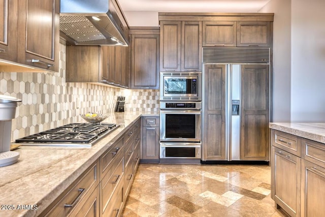 kitchen featuring light stone counters, decorative backsplash, built in appliances, and wall chimney exhaust hood