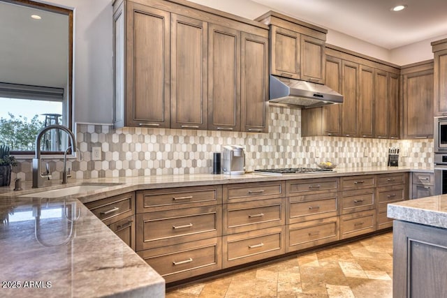 kitchen featuring under cabinet range hood, a sink, stainless steel appliances, decorative backsplash, and light stone countertops