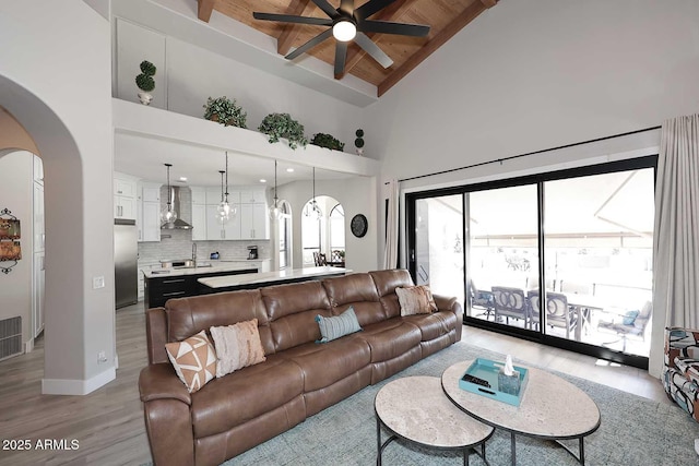 living area with beamed ceiling, light wood-style flooring, arched walkways, high vaulted ceiling, and a ceiling fan