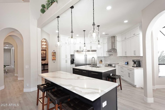 kitchen featuring a kitchen island with sink, appliances with stainless steel finishes, arched walkways, wall chimney exhaust hood, and a sink