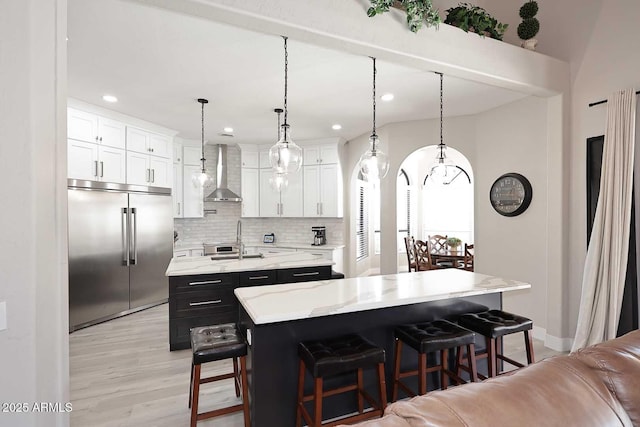 kitchen featuring wall chimney range hood, a center island with sink, stainless steel built in fridge, decorative backsplash, and light wood-style floors