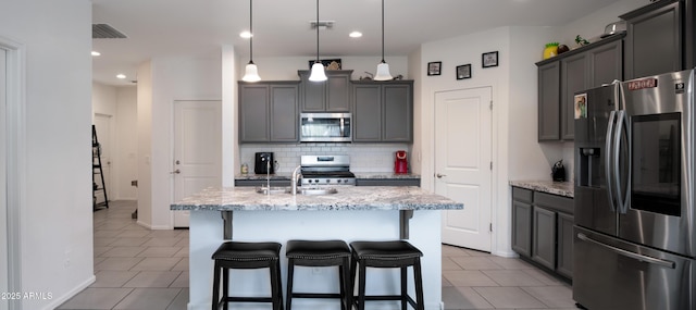 kitchen featuring stainless steel appliances, a kitchen breakfast bar, an island with sink, backsplash, and light stone countertops