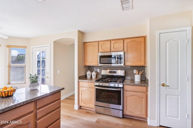 kitchen with appliances with stainless steel finishes, light brown cabinetry, tasteful backsplash, dark stone counters, and light hardwood / wood-style floors