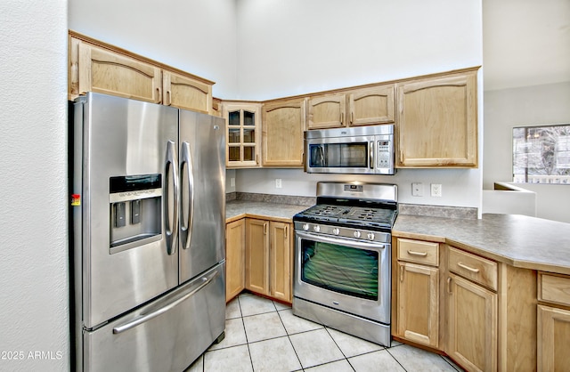 kitchen featuring light tile patterned floors, a peninsula, light brown cabinetry, stainless steel appliances, and glass insert cabinets