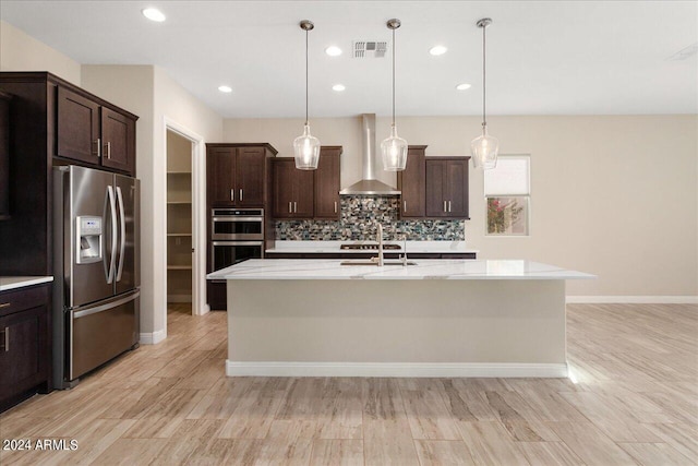 kitchen with hanging light fixtures, dark brown cabinets, a kitchen island with sink, wall chimney range hood, and appliances with stainless steel finishes