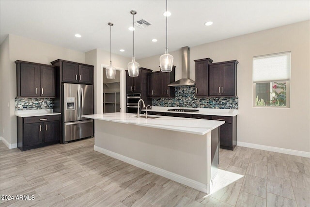 kitchen with wall chimney exhaust hood, backsplash, a center island with sink, appliances with stainless steel finishes, and decorative light fixtures