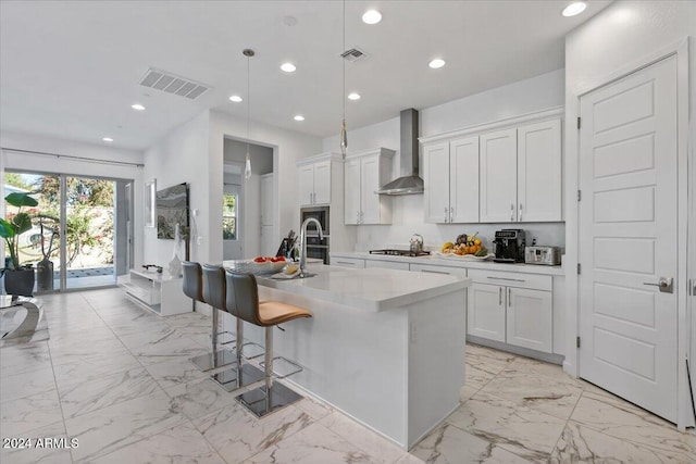 kitchen featuring wall chimney range hood, pendant lighting, a center island with sink, white cabinets, and stainless steel gas stovetop