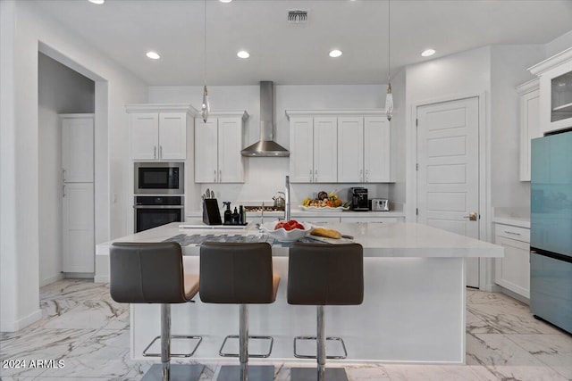 kitchen featuring wall chimney exhaust hood, white cabinetry, a kitchen island with sink, and appliances with stainless steel finishes