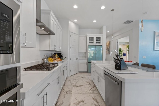 kitchen featuring wall chimney range hood, an island with sink, decorative light fixtures, white cabinets, and appliances with stainless steel finishes