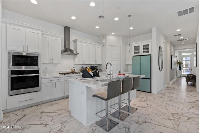 kitchen featuring a center island with sink, wall chimney range hood, hanging light fixtures, appliances with stainless steel finishes, and white cabinetry