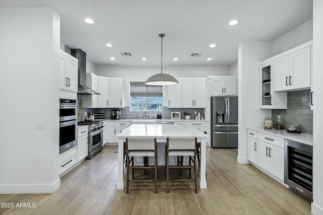 kitchen featuring wall chimney range hood, beverage cooler, visible vents, appliances with stainless steel finishes, and a kitchen breakfast bar