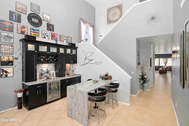 bar with a towering ceiling, beverage cooler, light tile patterned floors, and light stone counters