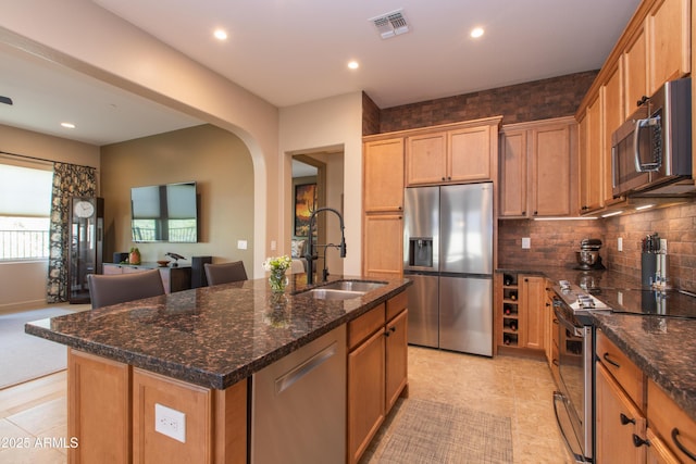 kitchen with stainless steel appliances, dark stone counters, decorative backsplash, sink, and a center island with sink