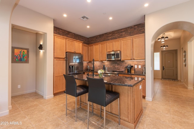 kitchen with backsplash, a kitchen bar, an island with sink, stainless steel appliances, and dark stone counters