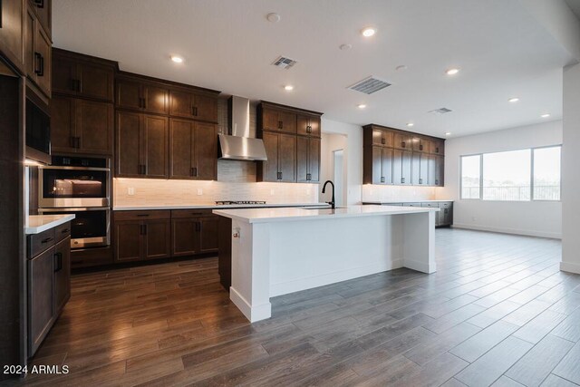 kitchen with gas cooktop, an island with sink, wall chimney range hood, and decorative backsplash