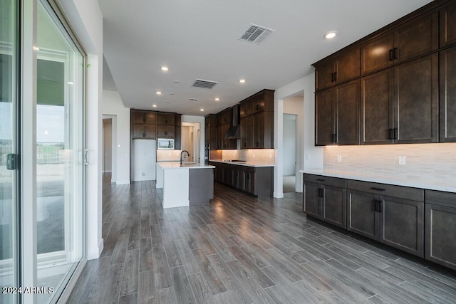 kitchen with sink, stainless steel microwave, wall chimney exhaust hood, an island with sink, and dark brown cabinets