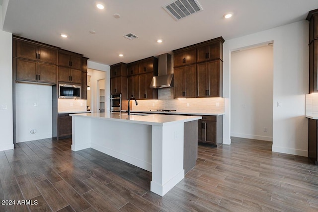 kitchen featuring built in microwave, a center island with sink, wall chimney range hood, sink, and oven