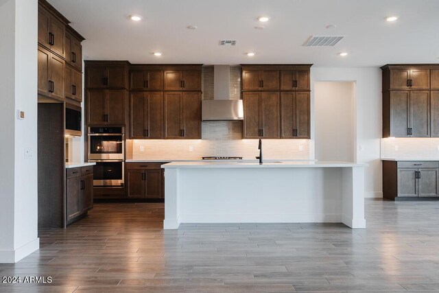 kitchen with light hardwood / wood-style flooring, backsplash, wall chimney range hood, an island with sink, and dark brown cabinetry