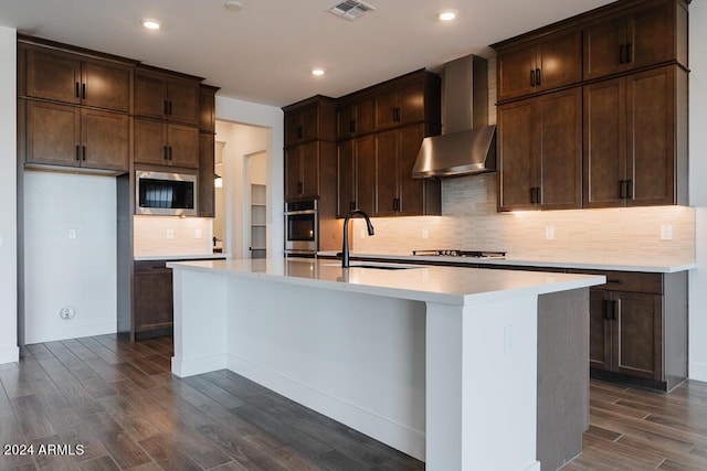 kitchen featuring an island with sink, sink, dark wood-type flooring, appliances with stainless steel finishes, and wall chimney exhaust hood