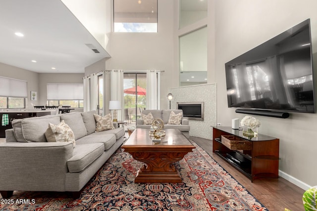 living area featuring a glass covered fireplace, a high ceiling, baseboards, and wood finished floors