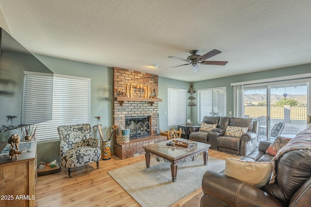 living room with ceiling fan, light wood-type flooring, a brick fireplace, and a textured ceiling