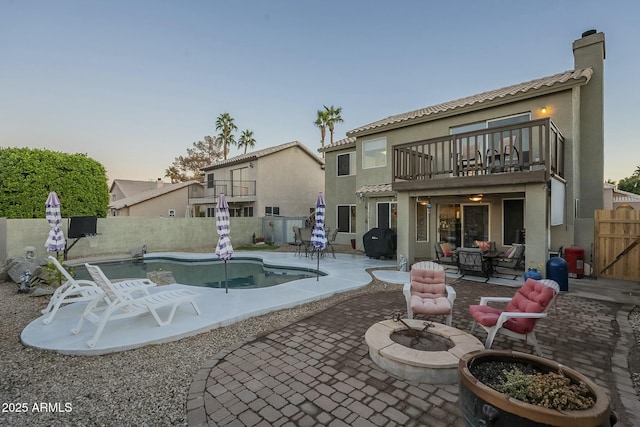 back house at dusk featuring an outdoor fire pit, a patio area, a balcony, and a fenced in pool