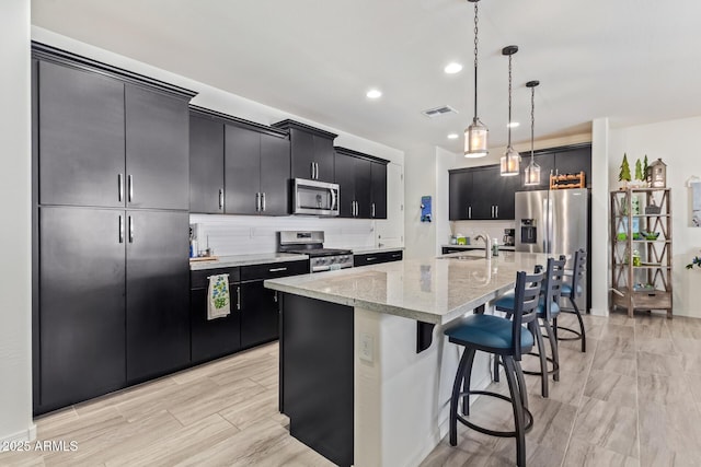 kitchen featuring pendant lighting, a center island with sink, stainless steel appliances, dark cabinets, and a kitchen breakfast bar