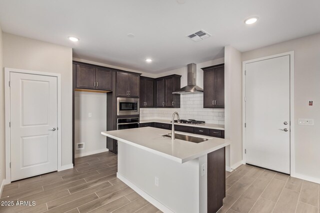 kitchen featuring wall chimney range hood, stainless steel appliances, tasteful backsplash, a kitchen island with sink, and dark brown cabinets