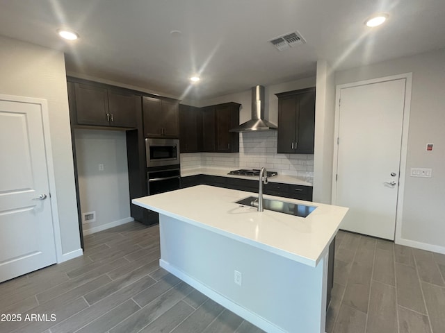 kitchen featuring a center island with sink, sink, wall chimney exhaust hood, decorative backsplash, and stainless steel appliances