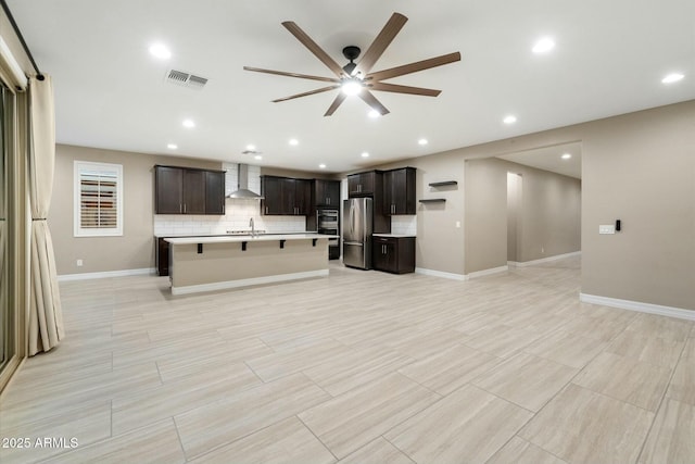 kitchen with an island with sink, stainless steel fridge, sink, tasteful backsplash, and wall chimney range hood