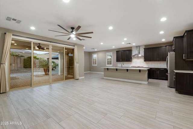 kitchen with an island with sink, wall chimney range hood, backsplash, stainless steel fridge, and a kitchen bar