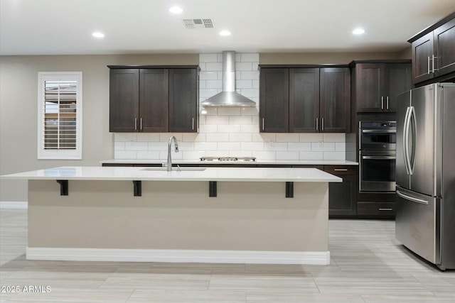 kitchen with appliances with stainless steel finishes, sink, a kitchen breakfast bar, wall chimney exhaust hood, and an island with sink