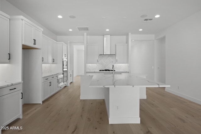 kitchen featuring an island with sink, wall chimney range hood, white cabinetry, and light stone countertops