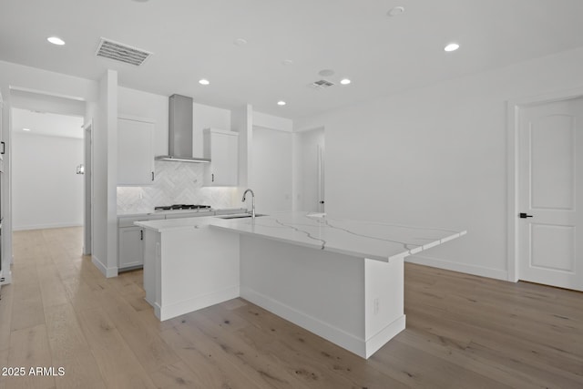 kitchen featuring light stone counters, a kitchen island with sink, wall chimney exhaust hood, and white cabinetry