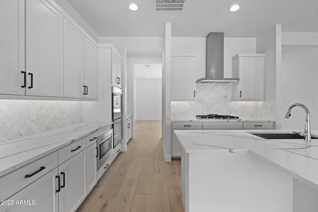 kitchen featuring white cabinets, wall chimney exhaust hood, light stone counters, stainless steel appliances, and a sink