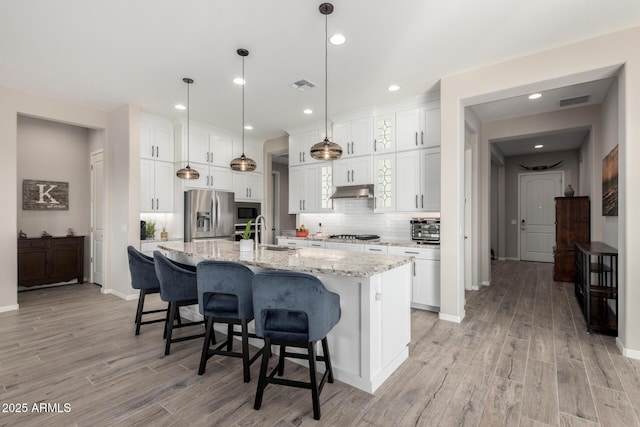 kitchen with light stone counters, stainless steel appliances, a center island with sink, white cabinetry, and hanging light fixtures