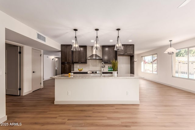 kitchen featuring decorative light fixtures, a kitchen island with sink, wall chimney exhaust hood, and dark brown cabinets