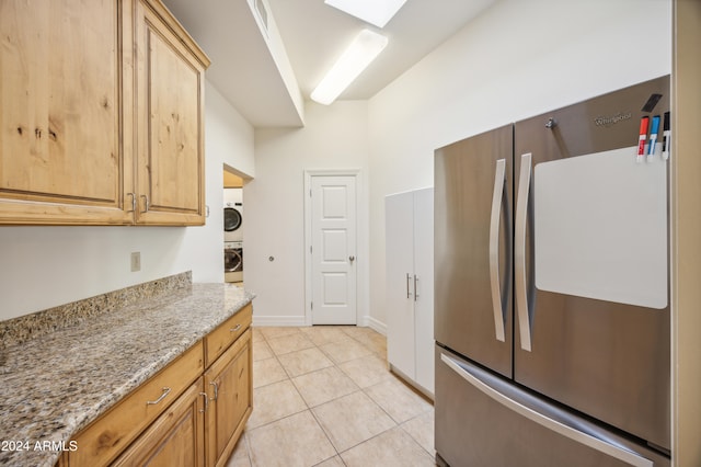 kitchen with light brown cabinets, stainless steel fridge, light tile patterned floors, light stone counters, and stacked washer / dryer