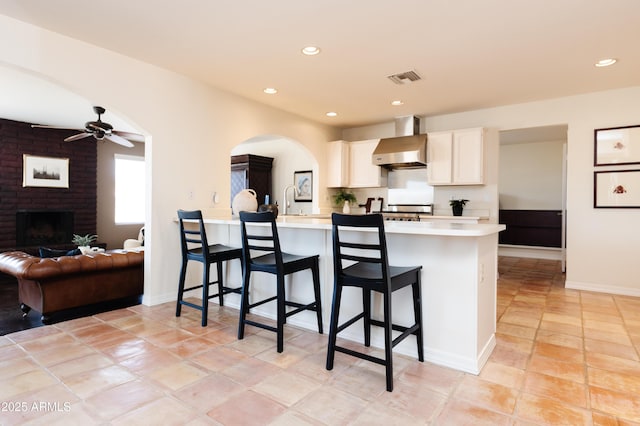 kitchen with a breakfast bar area, white cabinets, kitchen peninsula, a brick fireplace, and wall chimney range hood