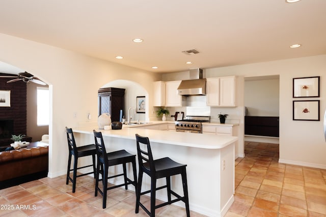 kitchen featuring a kitchen bar, sink, stainless steel range, kitchen peninsula, and wall chimney range hood