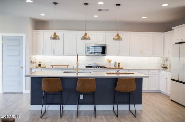 kitchen featuring stainless steel microwave, freestanding refrigerator, a kitchen island with sink, light countertops, and white cabinetry