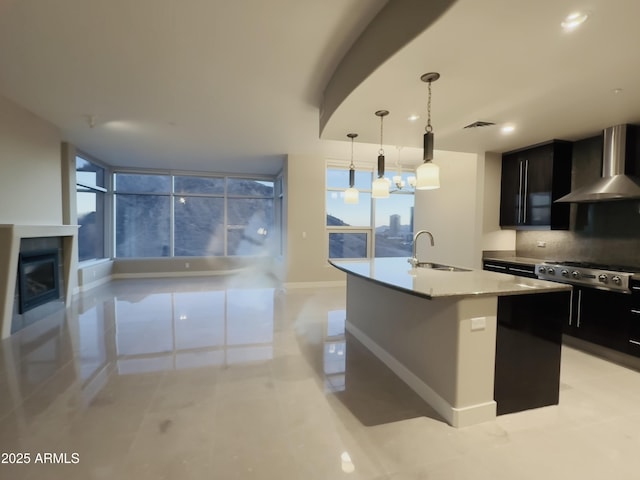 kitchen with stainless steel gas cooktop, sink, an island with sink, pendant lighting, and wall chimney range hood