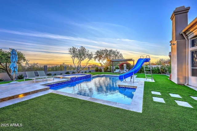 pool at dusk featuring an in ground hot tub, a patio, a water slide, and a yard